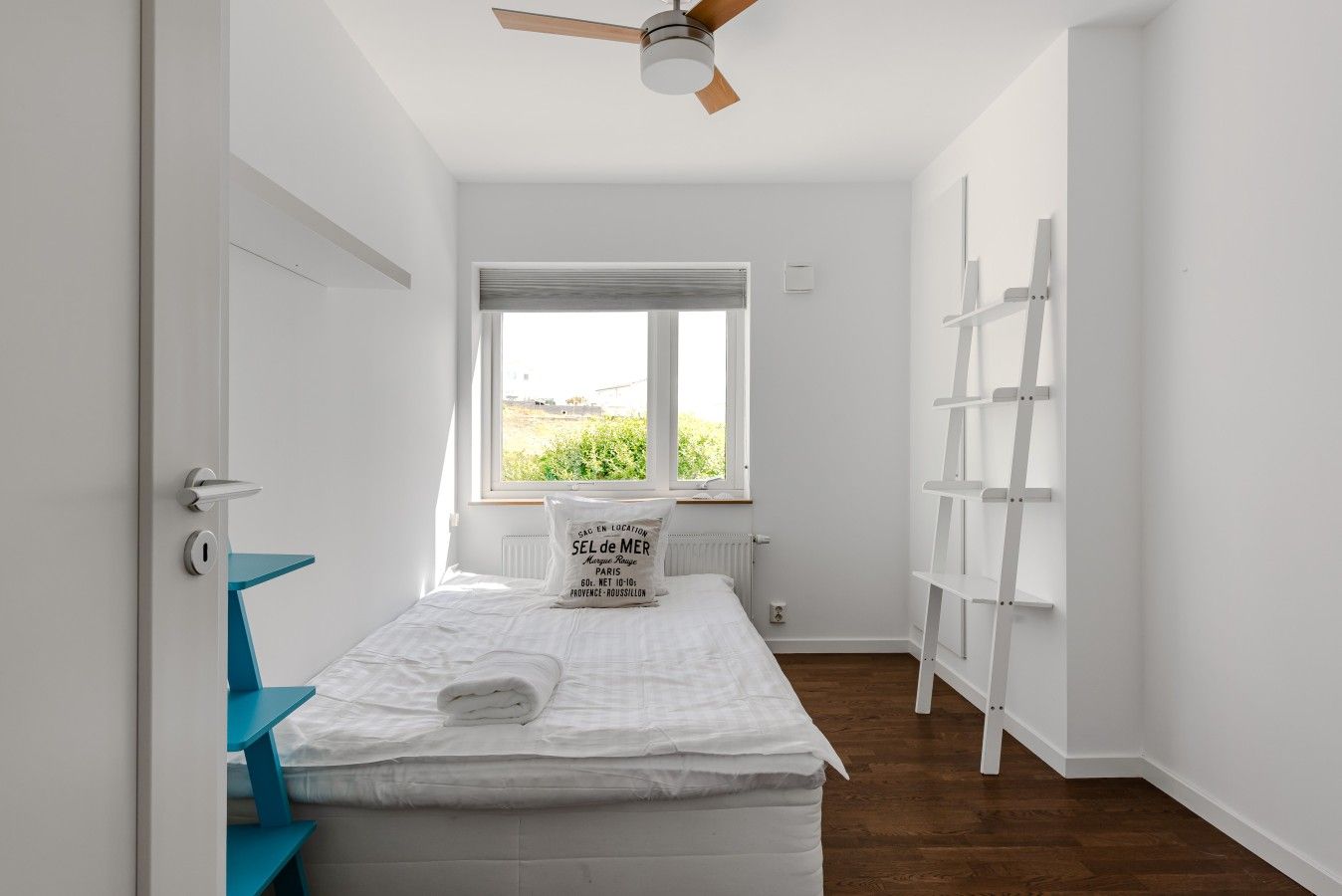 A bedroom with a bed, a window, a ceiling fan, and a white wall with a shelf.