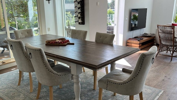 Uma sala de jantar com uma mesa de madeira, seis cadeiras estofadas, um tapete e uma televisão na parede.