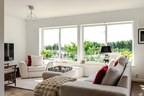 En moderne stue med sofa, sofabord og fjernsyn. Store vinduer med udsigt til grønne omgivelser.