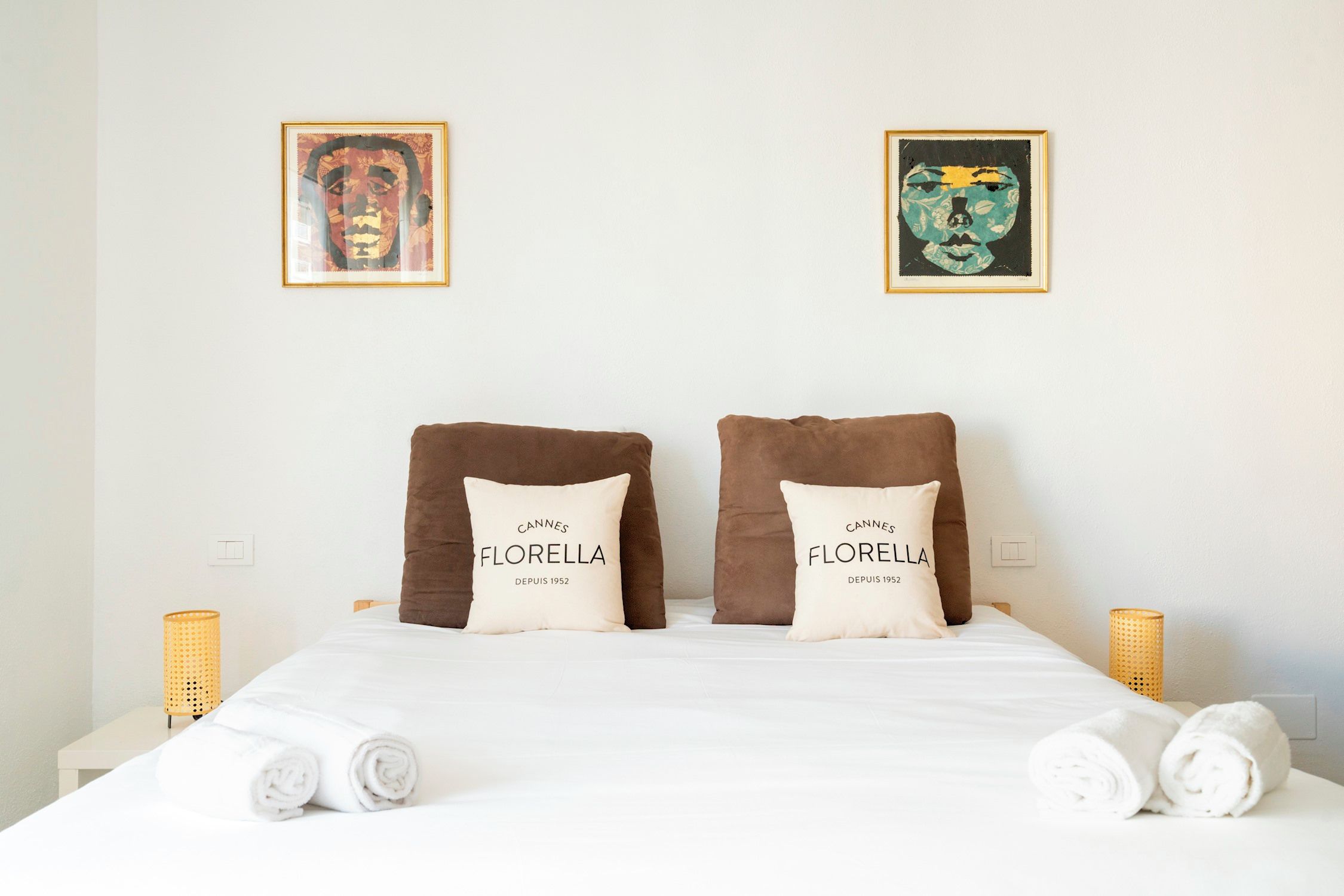 A bed with two pillows labeled 'CANNES FLORELLA DEPUIS 1952', two rolled towels, and two wall-mounted lamps.