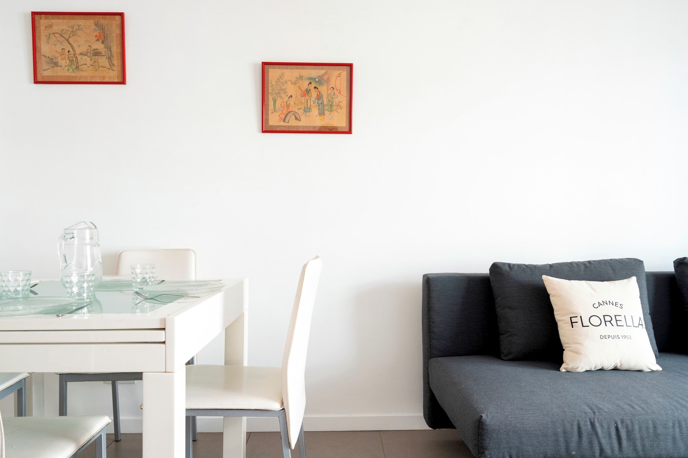 Un soggiorno minimalista con zona pranzo, caratterizzato da un divano con un cuscino con la scritta 'CANNES FLORELLA' e due opere d'arte incorniciate appese al muro.