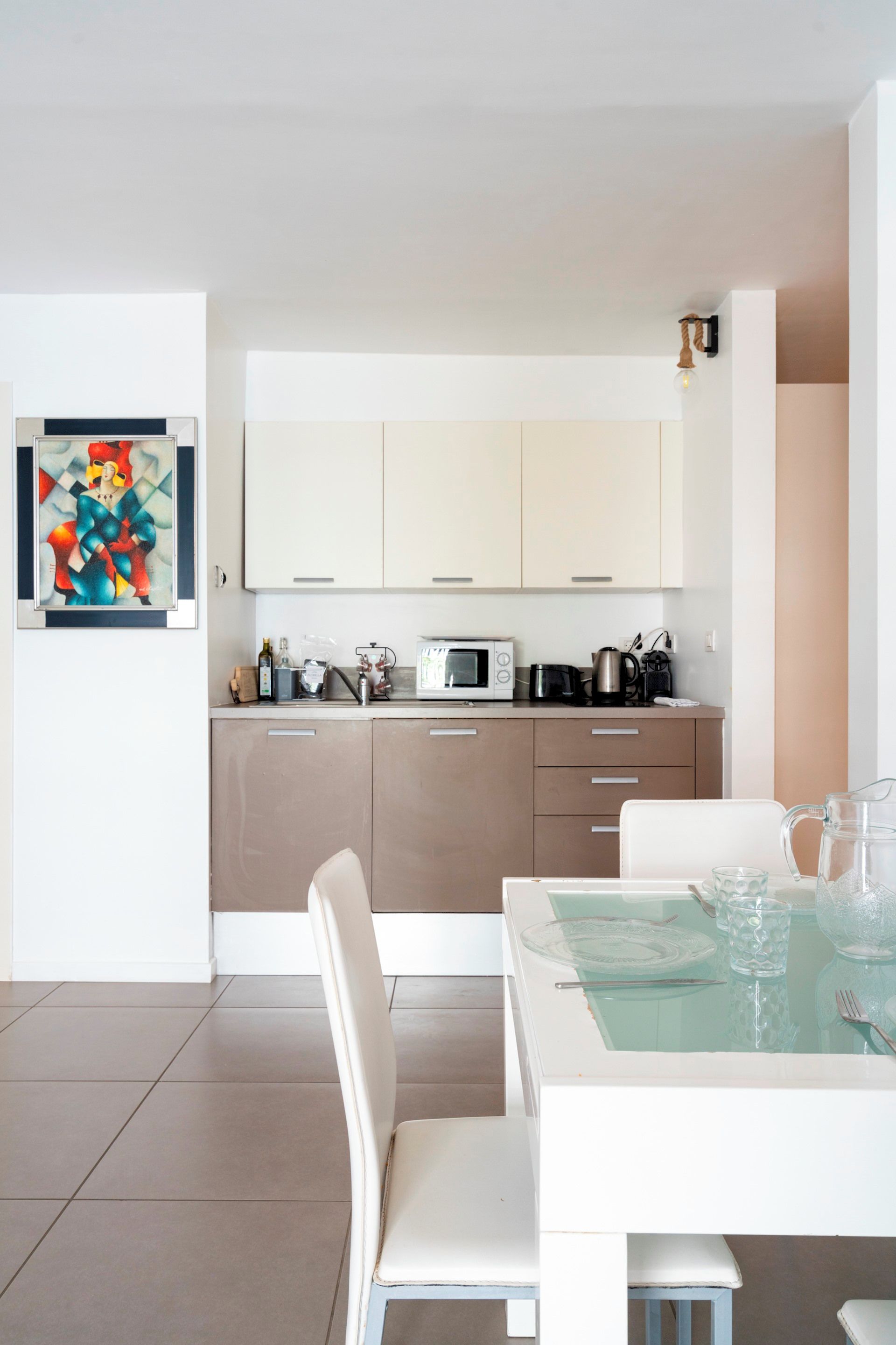 A modern kitchen with white cabinets, a dining table with chairs, and a glass-top table with a pitcher and glasses.