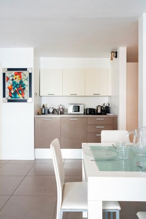 A modern kitchen with white cabinets, a dining table with chairs, and a glass-top table with a pitcher and glasses. - Le Palm Beach aperçu mer avec balcon proche plages (Cannes)