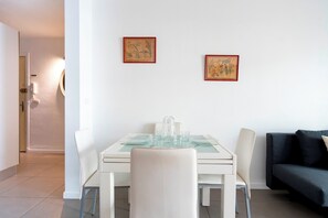 Ein Essbereich mit einem Tisch für zwei Personen, zwei gerahmten Kunstwerken an der Wand und einem dunkelgrauen Sofa im Hintergrund.