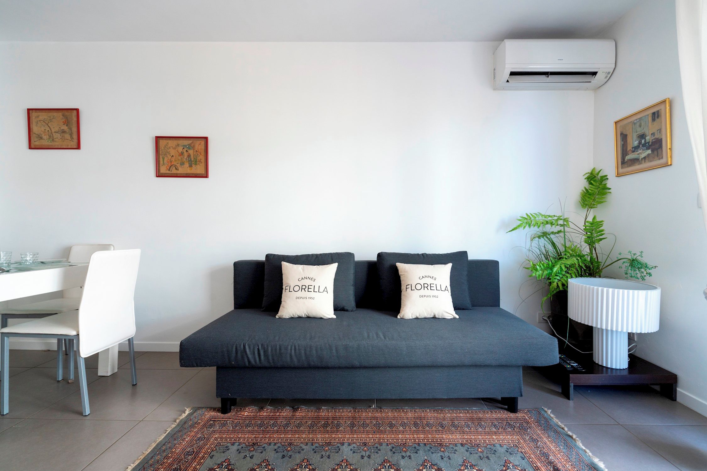 A modern living room with a sofa, two pillows, a dining table with chairs, a potted plant, and an air conditioning unit.