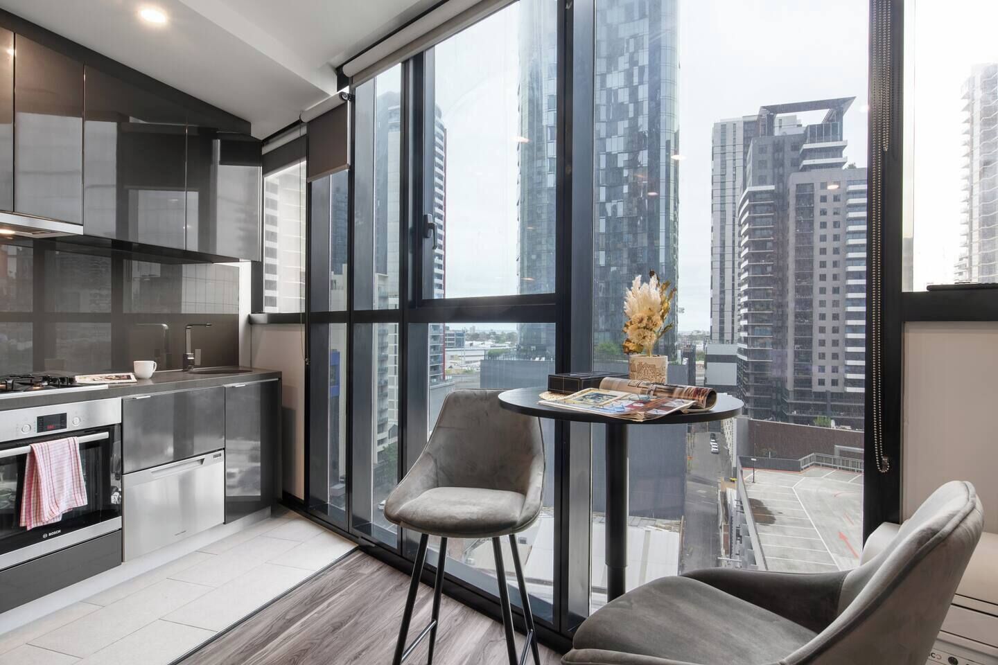 A modern kitchen with stainless steel appliances, a dining area with a round table and two chairs, and a large window offering a city view.