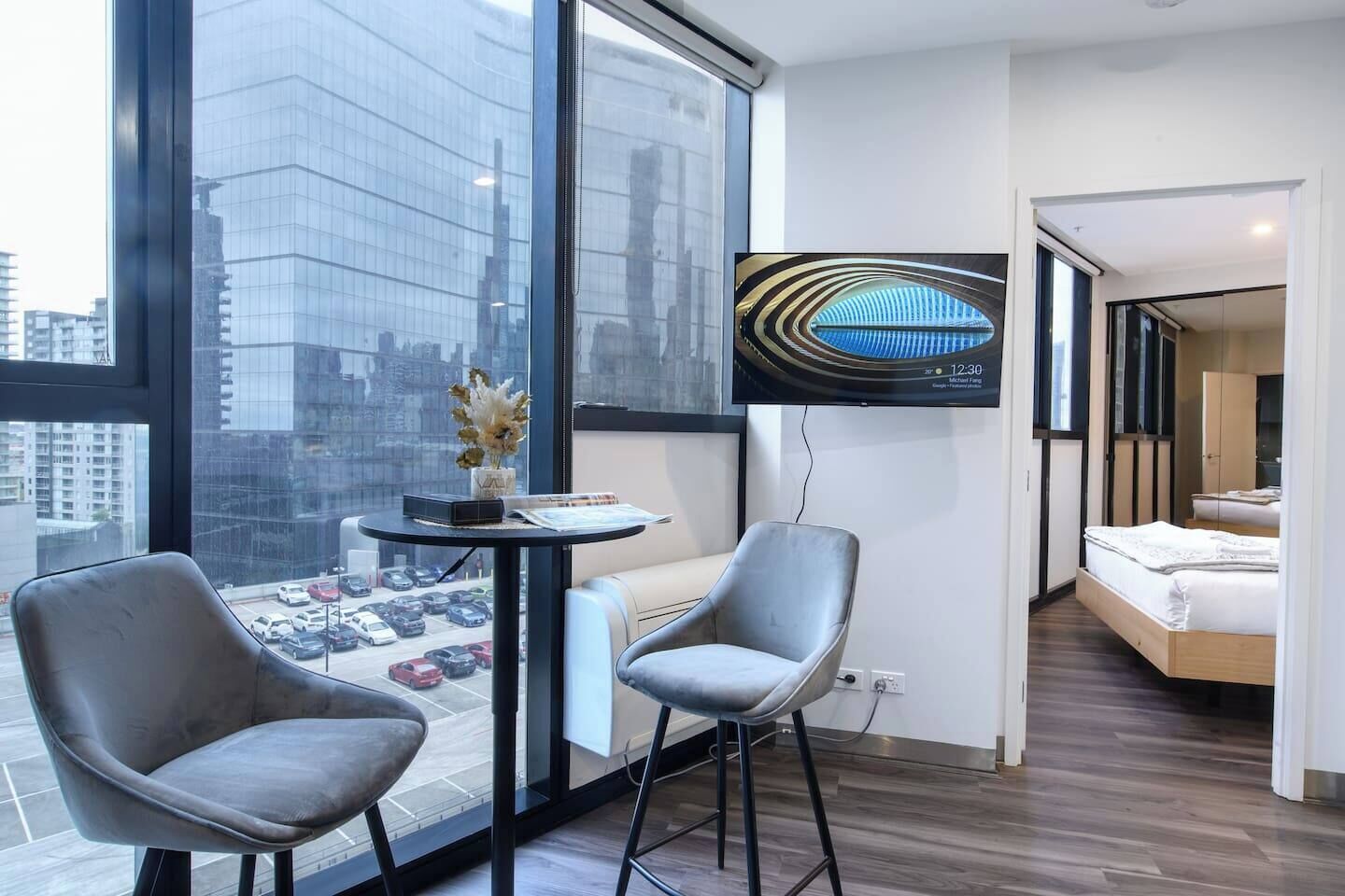 A modern hotel room with a large window, a flat-screen TV, a small table with a vase, two chairs, and a bed.