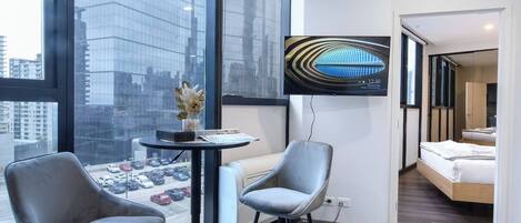 A modern hotel room with a large window, a flat-screen TV, a small table with a vase, two chairs, and a bed.