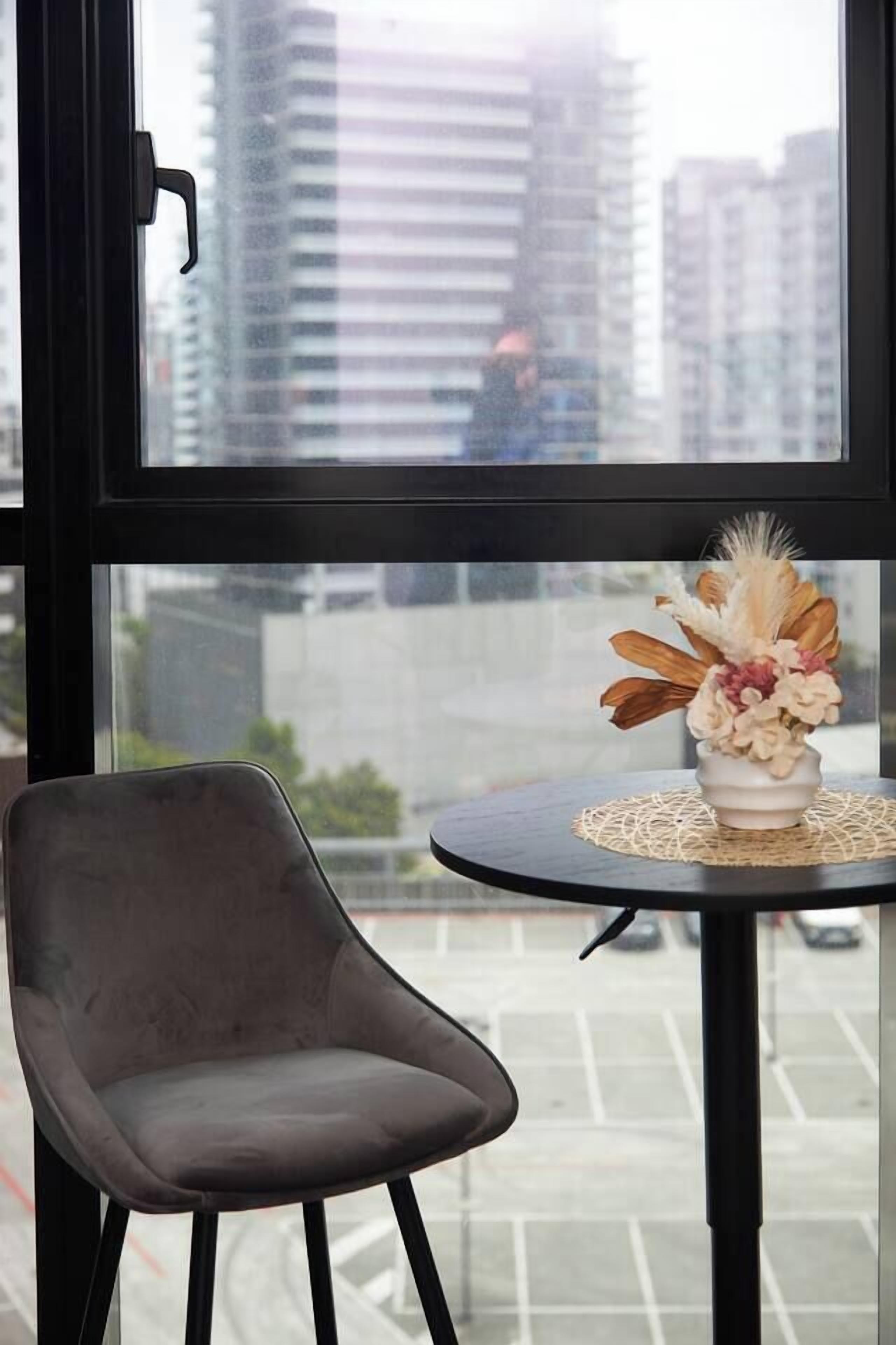 Une chambre moderne donnant sur un paysage urbain grâce à une grande fenêtre, avec une table ronde ornée d’un vase de fleurs et une seule chaise.