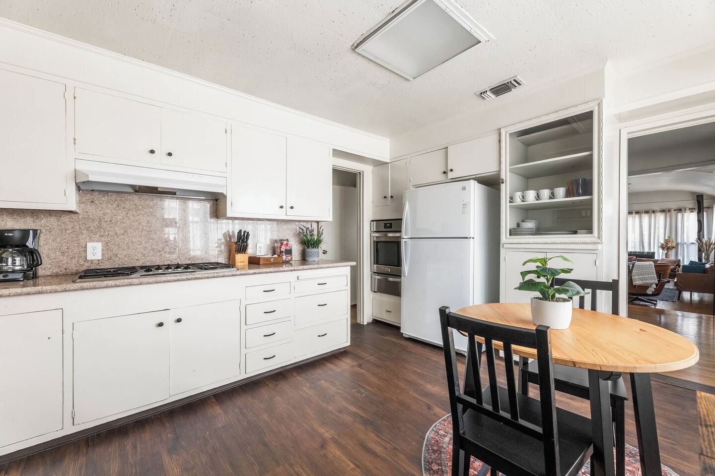 Een moderne keuken met witte kasten, een ingebouwde oven en een koelkast. Er is een eethoek met een houten tafel en stoelen.