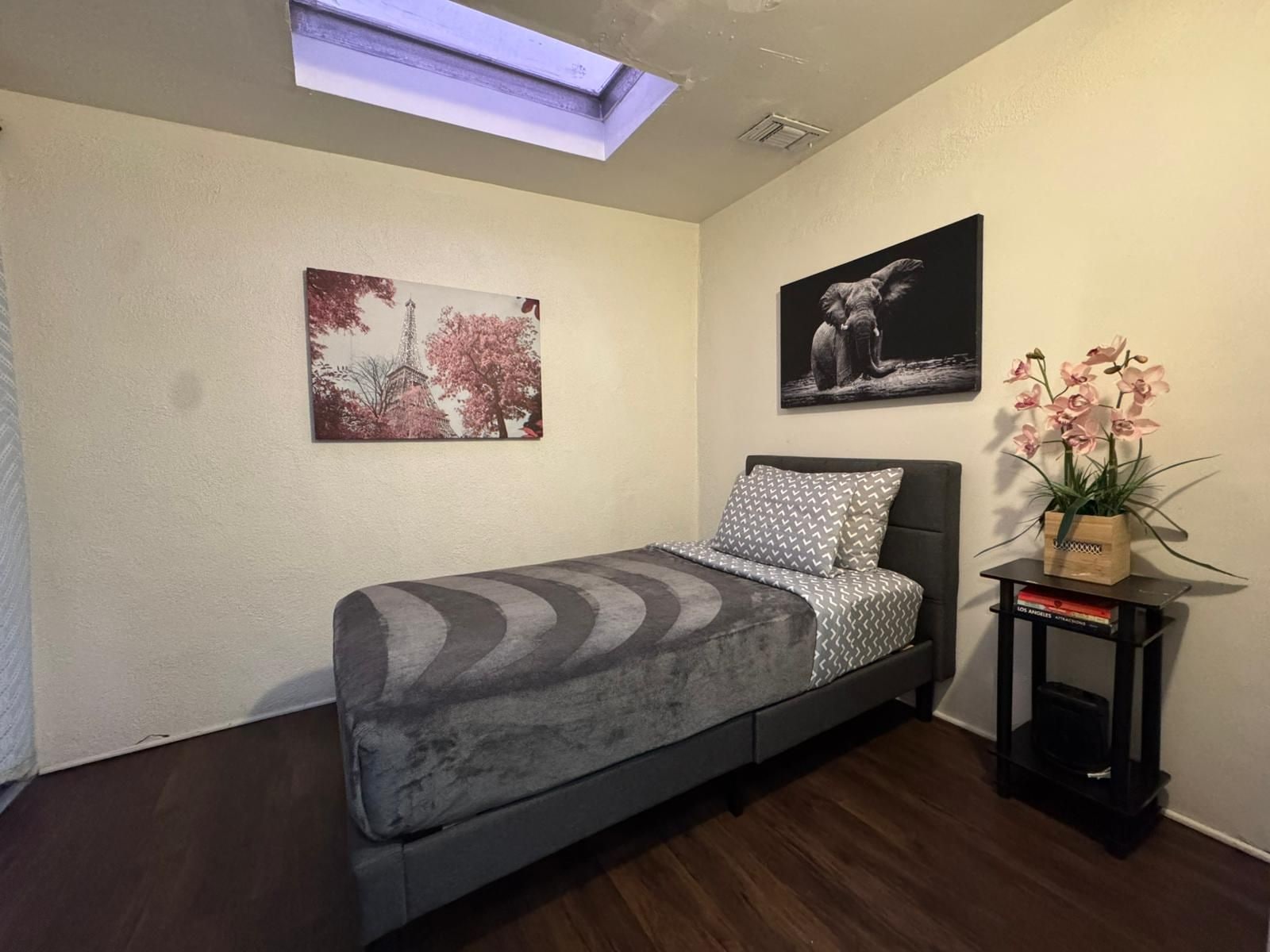 Une chambre à coucher avec un lit, une lucarne, une petite table avec une plante et deux tableaux encadrés accrochés au mur.