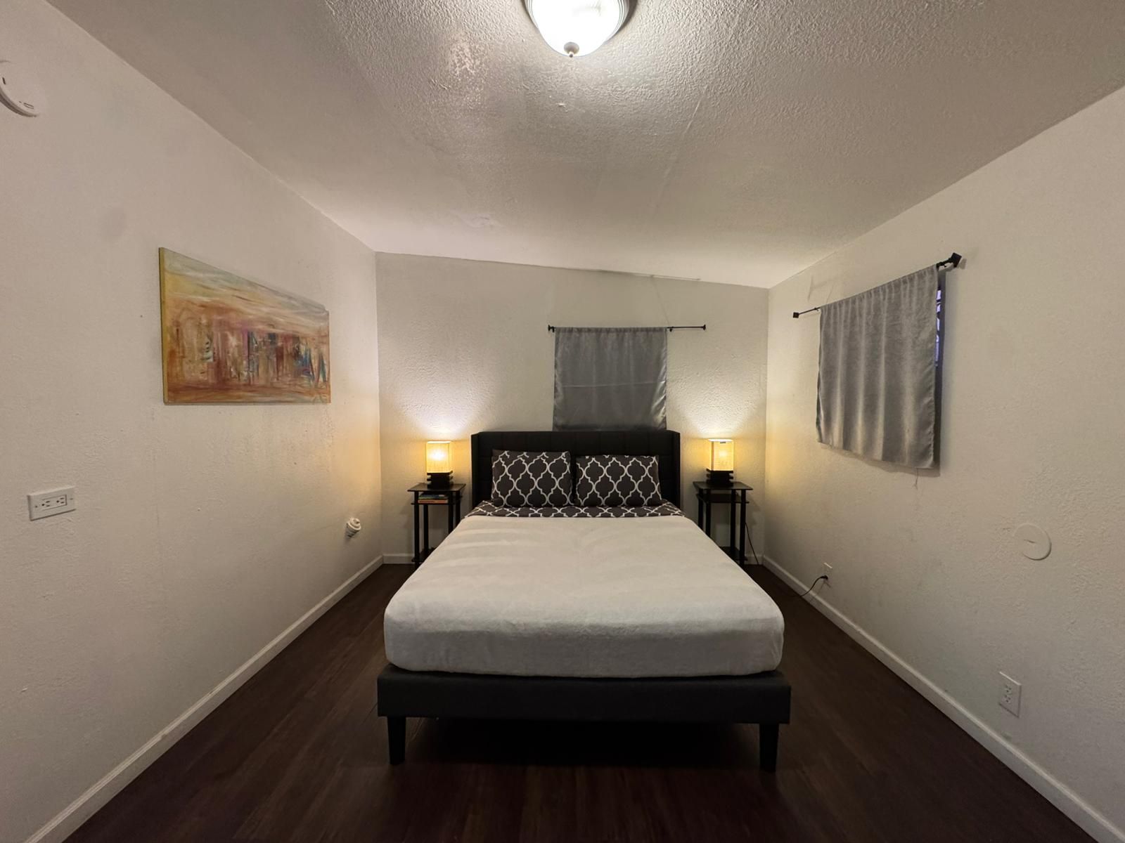 A bedroom with a bed, two bedside lamps, a painting on the wall, and a window with curtains.