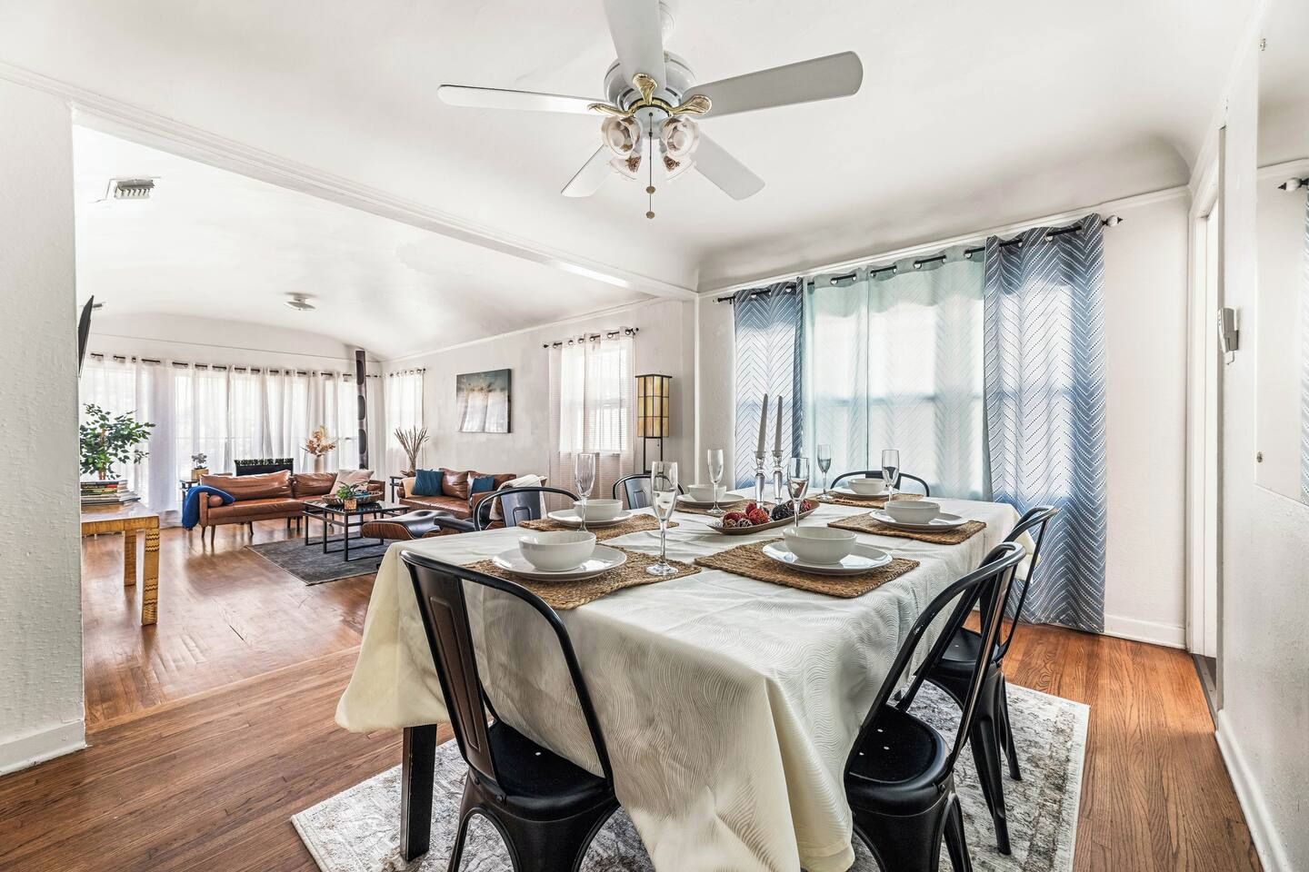 Un coin repas avec une table dressée pour quatre personnes, un ventilateur de plafond et une vue sur un salon comprenant un canapé et une table basse.