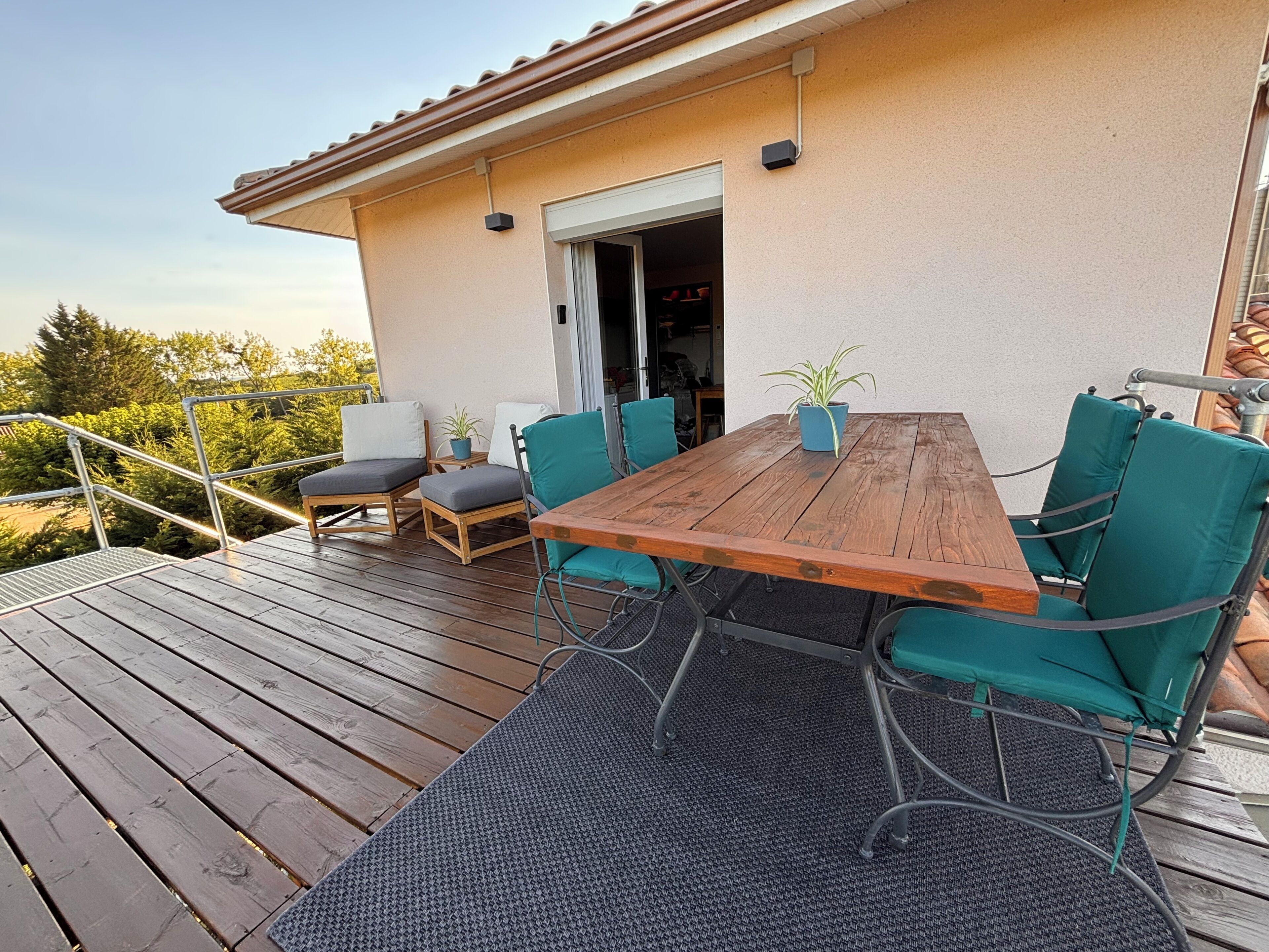 Una terraza de madera con una mesa y sillas, una planta en maceta y una puerta que da a un espacio interior.