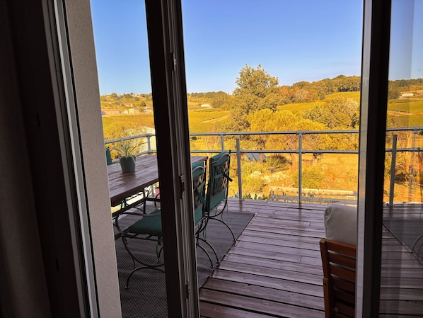 A balcony with a wooden floor, a table, and chairs, overlooking a rural landscape with trees and fields.
