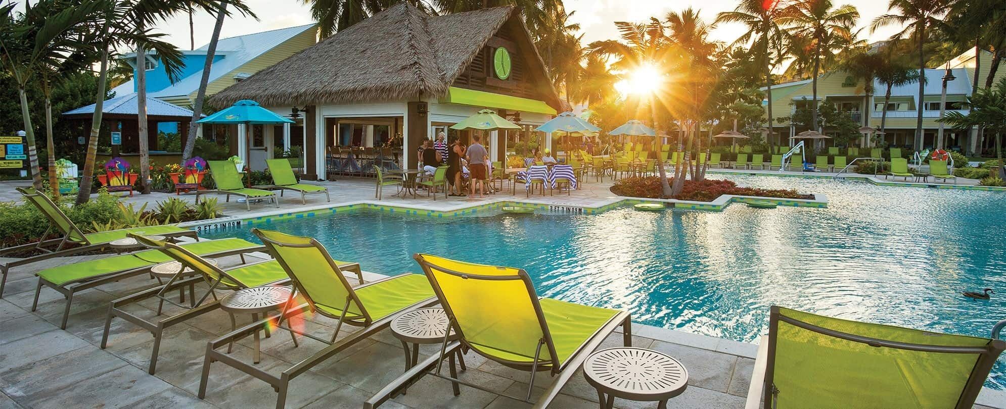 A resort pool area with lounge chairs, a thatched-roof building, and a clear view of the sunset.