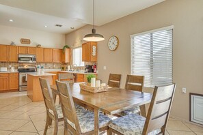 A kitchen with wooden cabinets, a dining table with chairs, a clock, and a window with blinds. - The Surprise Oasis, Heated Pool, Pet Friendly (Surprise)