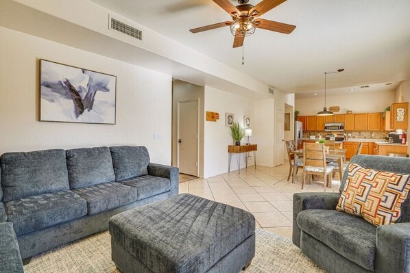A living room with a grey sofa, a matching ottoman, and a patterned armchair. There is a ceiling fan, a dining area with a table and chairs, and a kitchen in the background. - The Surprise Oasis, Heated Pool, Pet Friendly (Surprise)