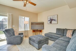 A living room with a ceiling fan, a flat-screen TV, and a large window with blinds. - The Surprise Oasis, Heated Pool, Pet Friendly (Surprise)