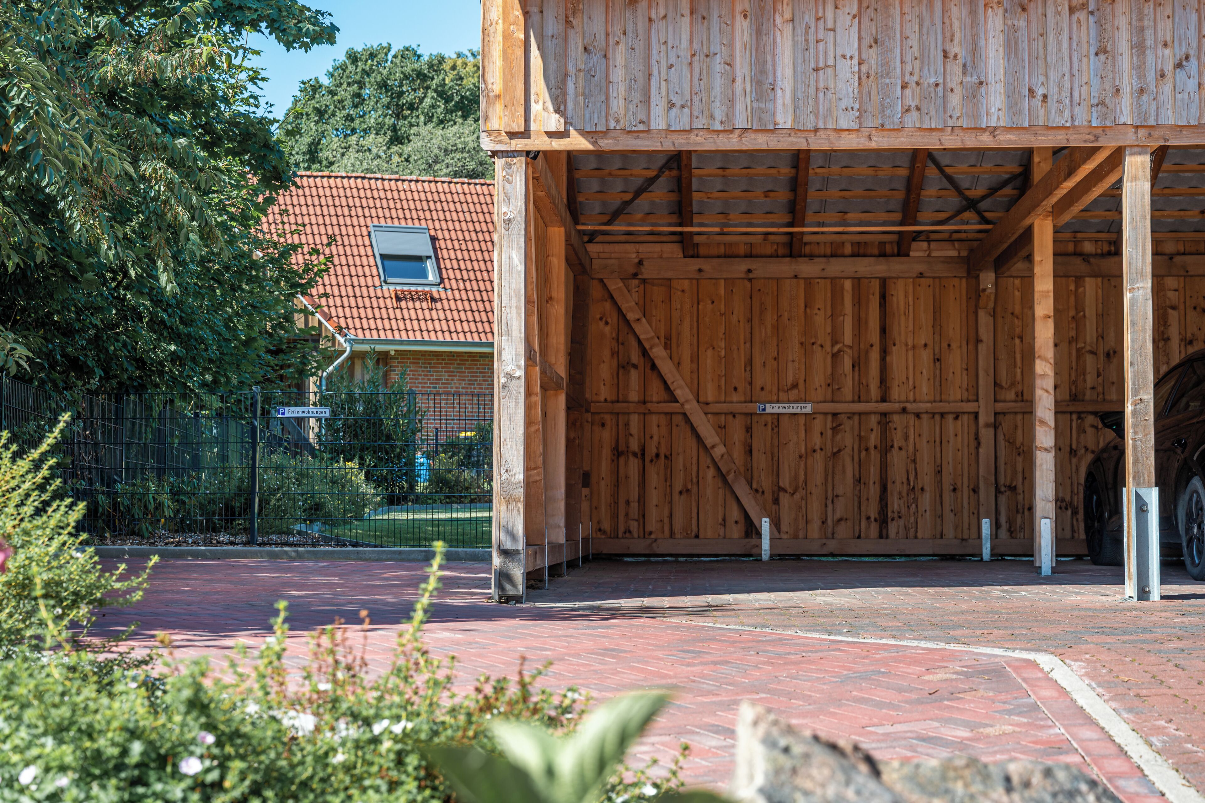 A wooden building with a large open garage space, a brick pathway, and a fenced area with greenery.