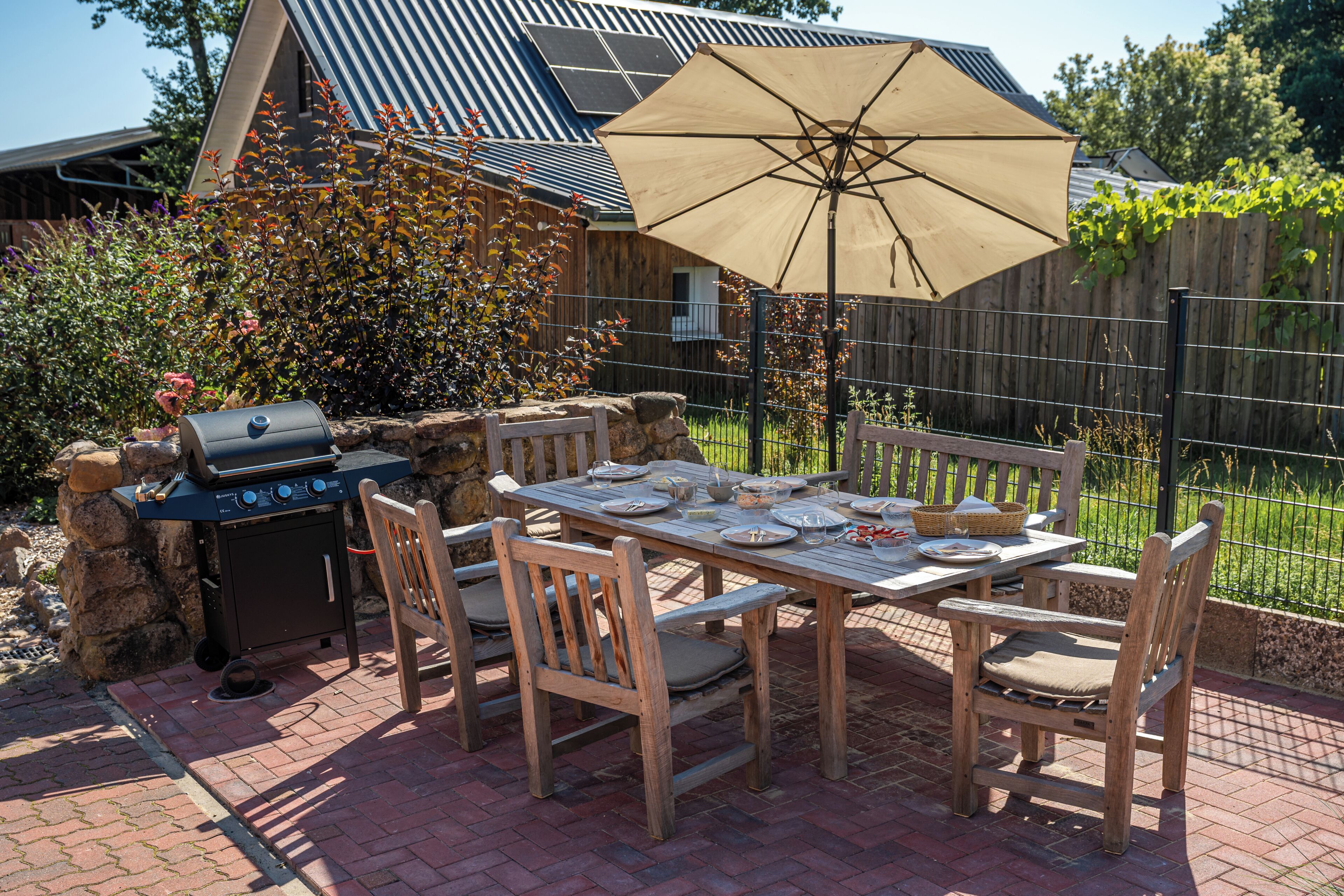 Eine Terrasse im Freien mit Esstisch, Stühlen, einem Grill und einem großen Sonnenschirm.