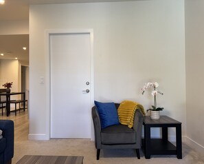 A room with a closed door, a grey armchair with a blue cushion, a small table with a potted orchid, and a bench with a yellow throw blanket. - Paradise in Glenridding (Edmonton)