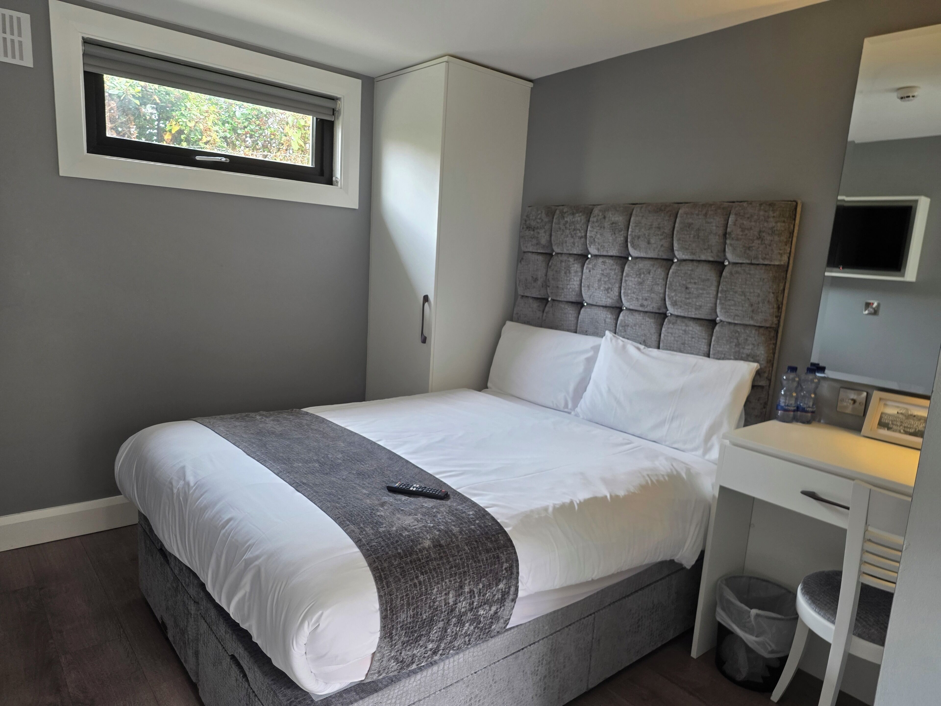 Un dormitorio con cama, cabecera, ventana, armario, escritorio y cubo de basura.