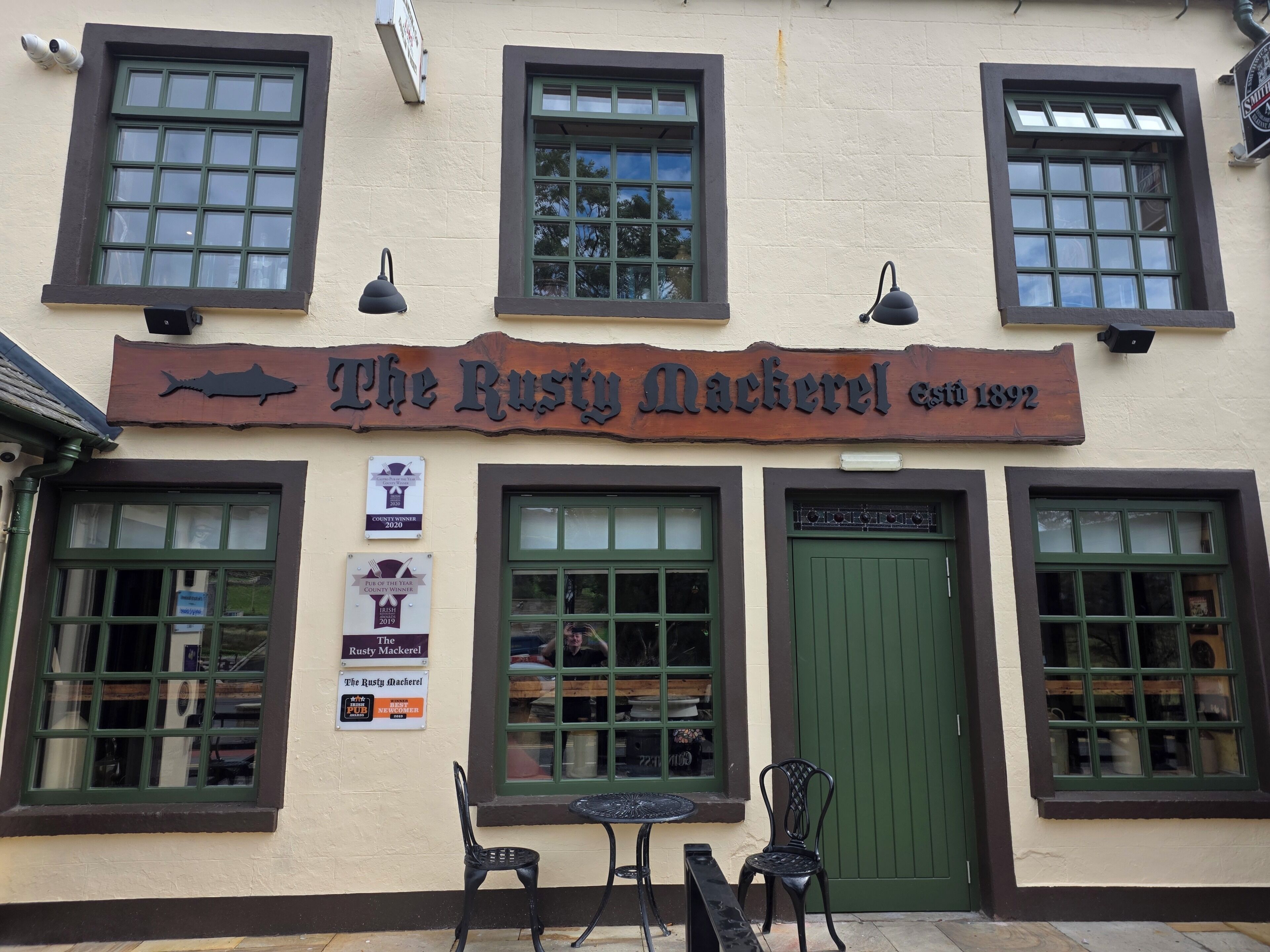 Um pub chamado 'The Rusty Mackerel', com fachada tradicional, várias janelas e área externa para sentar.