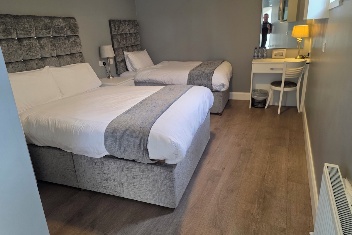 A hotel room with two beds, a desk, a chair, a lamp, and a radiator.