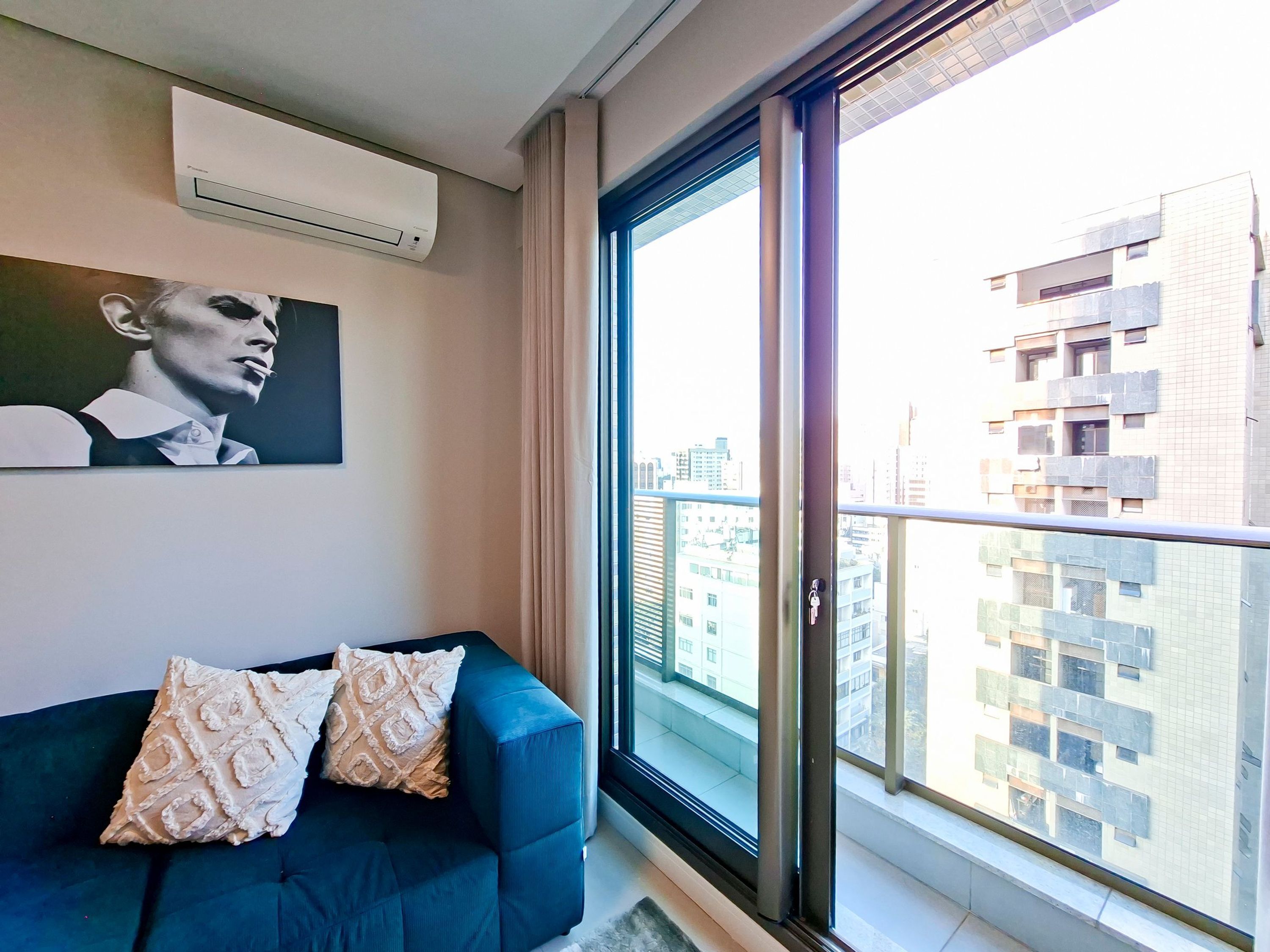 A modern living room with a blue sofa, patterned cushions, a mounted air conditioner, and a large window with a city view.