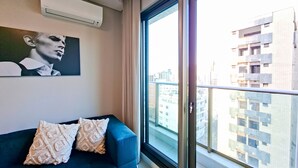 Un salon moderne avec un canapé bleu, des coussins à motifs, un climatiseur fixé au mur et une grande fenêtre donnant sur la ville.