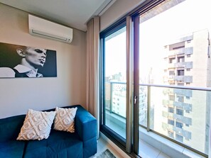 Ein modernes Wohnzimmer mit einem blauen Sofa, gemusterten Kissen, einer Wandklimaanlage und einem großen Fenster mit Blick auf die Stadt.