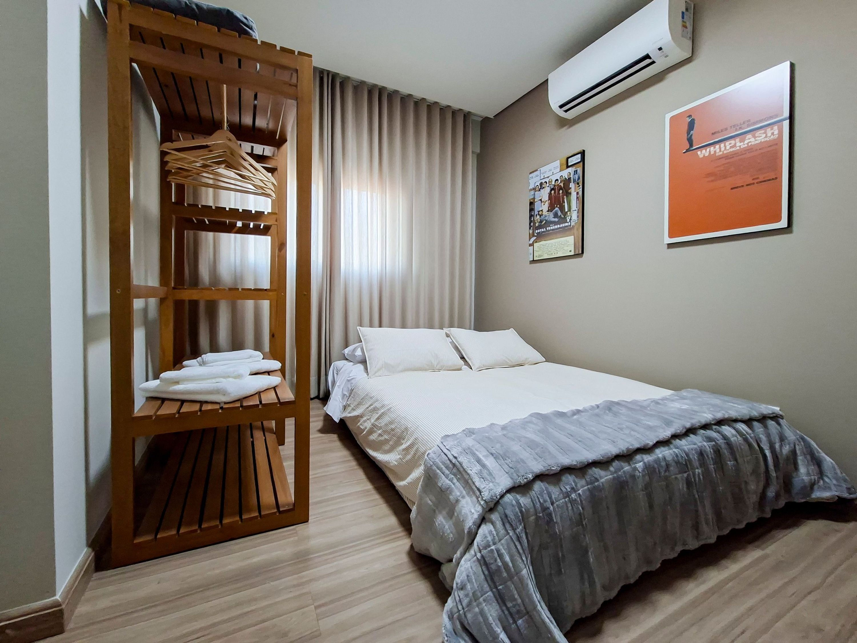 Quarto de hotel com uma cama, um guarda-roupa de madeira com roupas penduradas, um quadro na parede e um aparelho de ar condicionado.