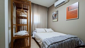 Une chambre d’hôtel avec un lit, une armoire en bois où sont suspendus des vêtements, une affiche au mur et un appareil de climatisation.