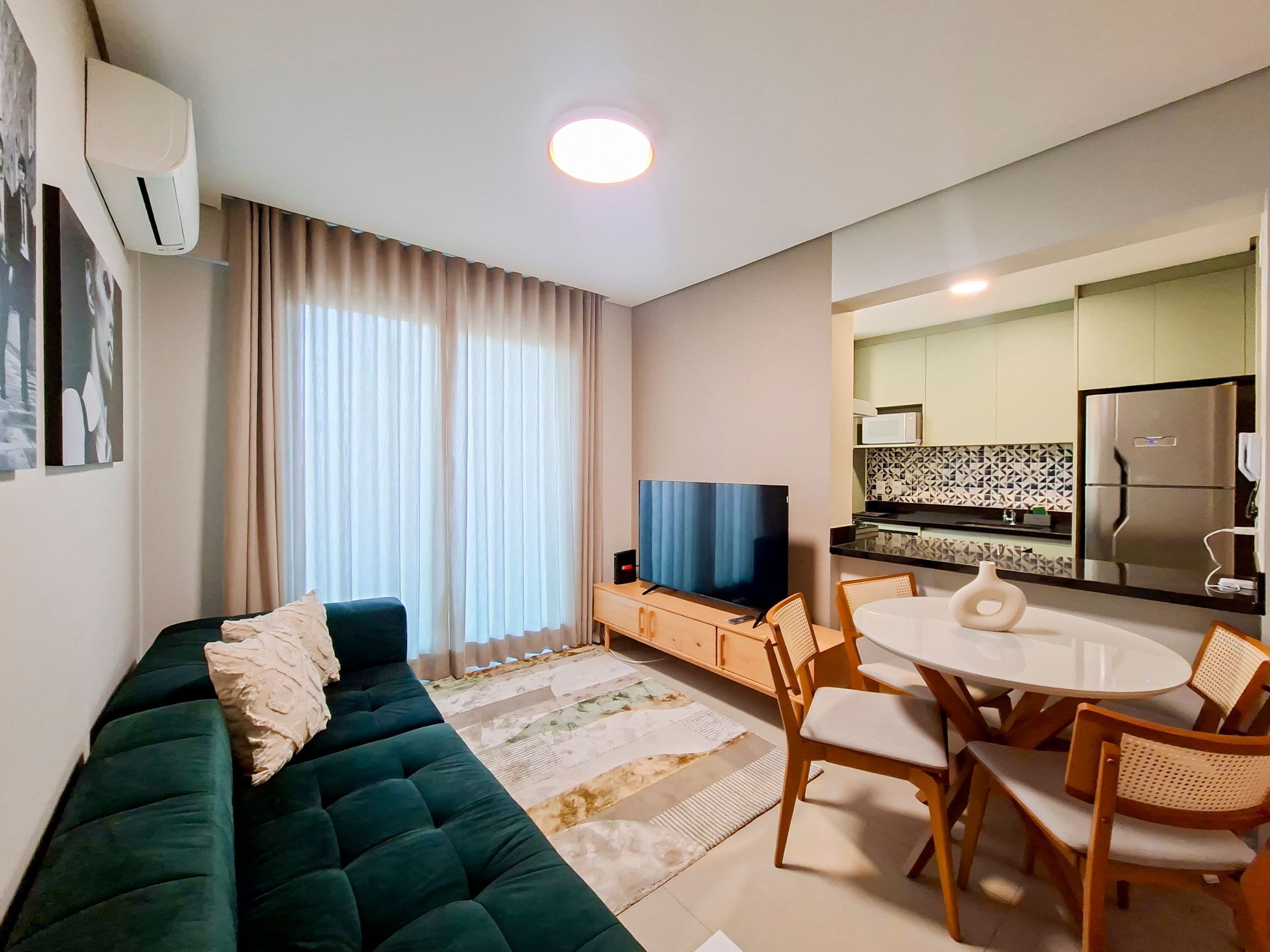 A modern living room with a green sofa, a flat-screen TV, a dining area with a round table and chairs, and a kitchen with a refrigerator and microwave.
