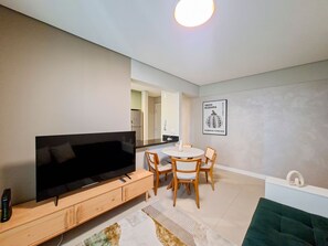 Ein modernes Wohnzimmer mit Flachbildfernseher, Holzmöbeln, einem Essbereich mit rundem Tisch und einer Küche im Hintergrund.