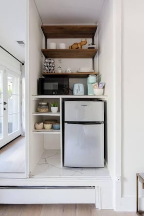 A compact kitchen with a built-in refrigerator, microwave, and toaster. There are shelves above the counter holding various kitchen items and a decorative animal figure.