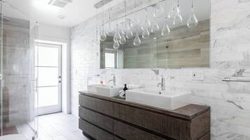 Un baño moderno con tocador doble, cabina de ducha de vidrio y un espejo grande con lámparas colgantes.