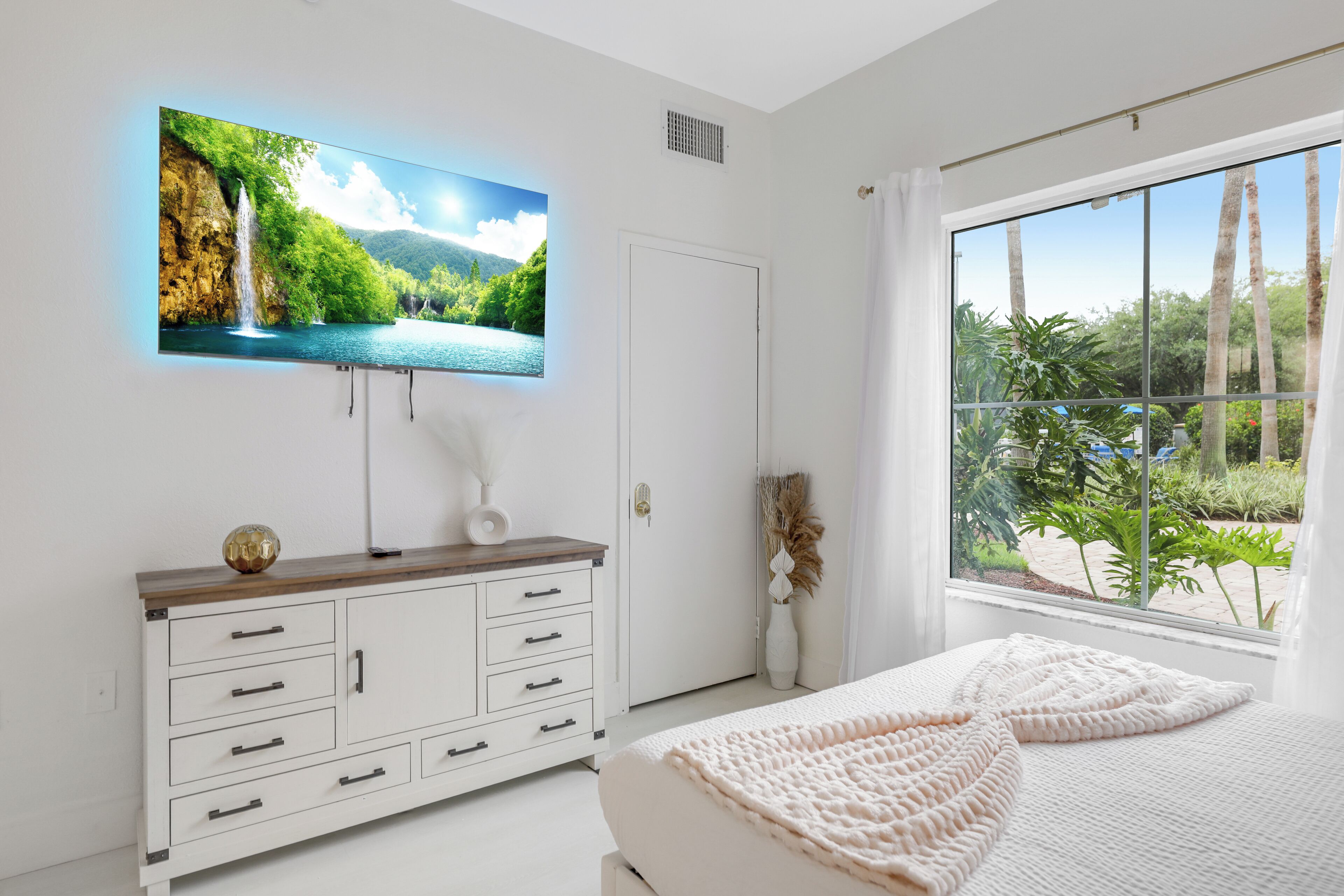 Une chambre à coucher avec un lit, un téléviseur fixé au mur, une fenêtre donnant sur un paysage verdoyant et une commode blanche avec des tiroirs.