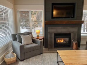Ein Wohnzimmer mit Kamin, einer Couch, einem Tisch und einem Fernseher, der über dem Kamin angebracht ist.