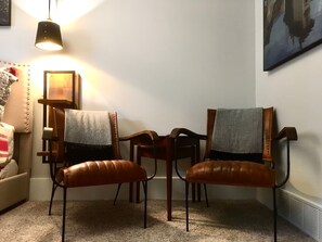 Ein Zimmer mit zwei Ledersesseln, einem kleinen Tisch, einer Couch, einer Pendelleuchte und einem gerahmten Bild an der Wand.