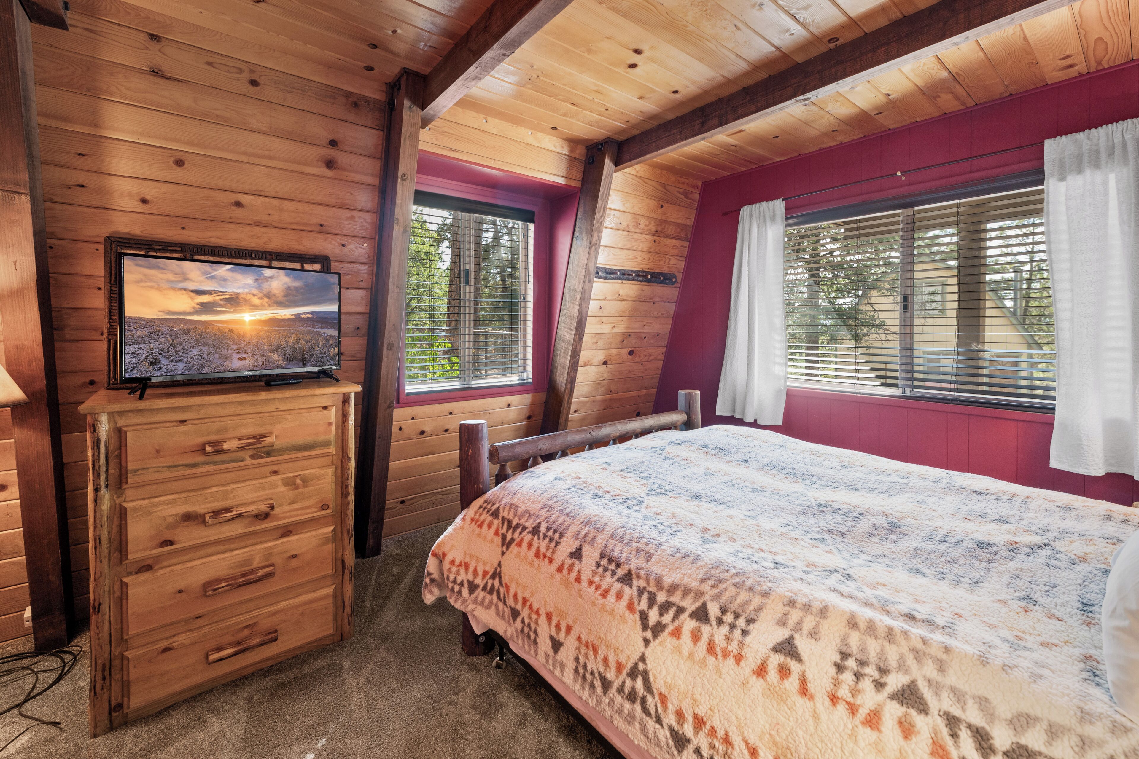 A wooden cabin room with a bed, a dresser, a television on the wall, and a window with blinds.
