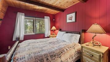 A bedroom with a wooden ceiling, a bed with a patterned blanket, two bedside tables with lamps, a window with curtains, and a framed picture on the wall.