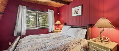 Quarto com teto de madeira, cama com manta estampada, duas mesas de cabeceira com luminárias, janela com cortinas e um quadro na parede.