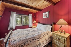 Un dormitorio con techo de madera, una cama con manta estampada, dos mesitas de noche con lámparas, una ventana con cortinas y un cuadro enmarcado en la pared.