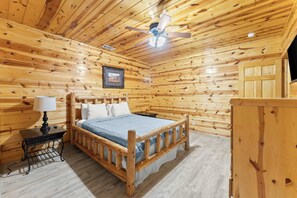 A wooden cabin room with a bed, a nightstand, a lamp, a ceiling fan, and a framed picture on the wall.