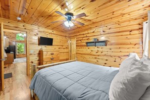 A wooden cabin bedroom with a bed, a ceiling fan, a television, and a window. - Inspiration by Blue Creek Cabins (Cleveland)