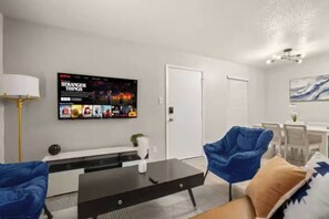 A modern living room with a flat-screen TV displaying 'Stranger Things', a black coffee table, blue armchairs, and a dining area with white furniture. - Central Austin • Parking • Sleeps 5 • Pet Friendly (Austin)