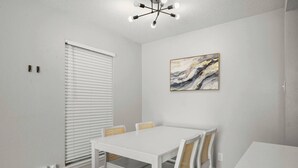 Ein Essbereich mit einem weißen Tisch und Stühlen, einer Deckenleuchte und einem an der Wand hängenden abstrakten Gemälde.