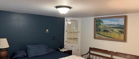 Ein Schlafzimmer mit Bett, Nachttisch, Lampe, einer Bank, einer Tür mit Milchglasfenster und einem Landschaftsbild an der Wand.